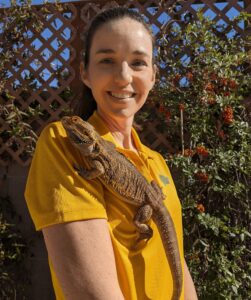 Photo of Lacey wearing her golden Leaping Lizards polo shirt and standing in front of a trellis with a bush growing up it. Lacey is holding Bailey the bearded dragon.